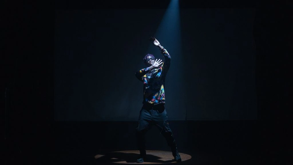A black-box student showcase with a performer on stage and an intimate student audience